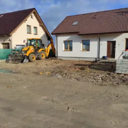 Żółta koparko-ładowarka JCB na placu budowy pomiędzy dwoma nowymi domami, widoczne palety z kostką brukową i błotniste podłoże.