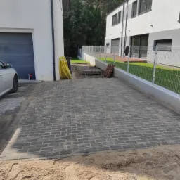 Ułożona szara kostka brukowa na podjeździe przed nowoczesnym domem, widoczne ślady piasku i żółte rury w tle.