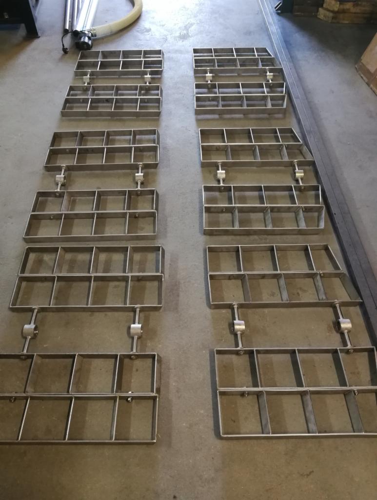 Metalowe ruszty spawane z profili kwadratowych, ułożone w dwóch rzędach na szarym betonowym podłożu, widoczne elementy instalacji w tle.