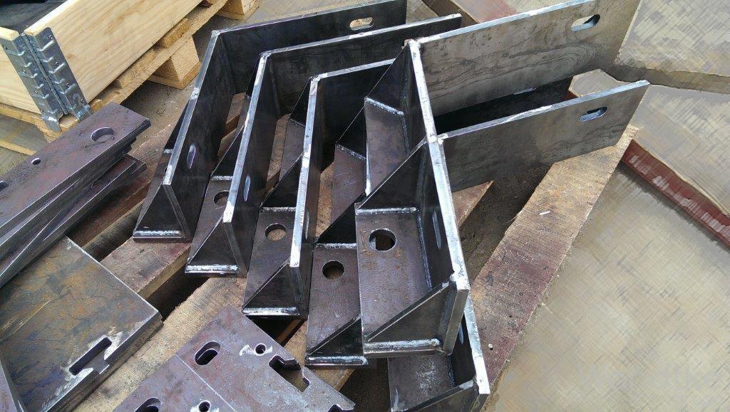 Metalowe, spawane wsporniki kątowe z otworami, ułożone na drewnianej palecie, widoczne ślady spawania.