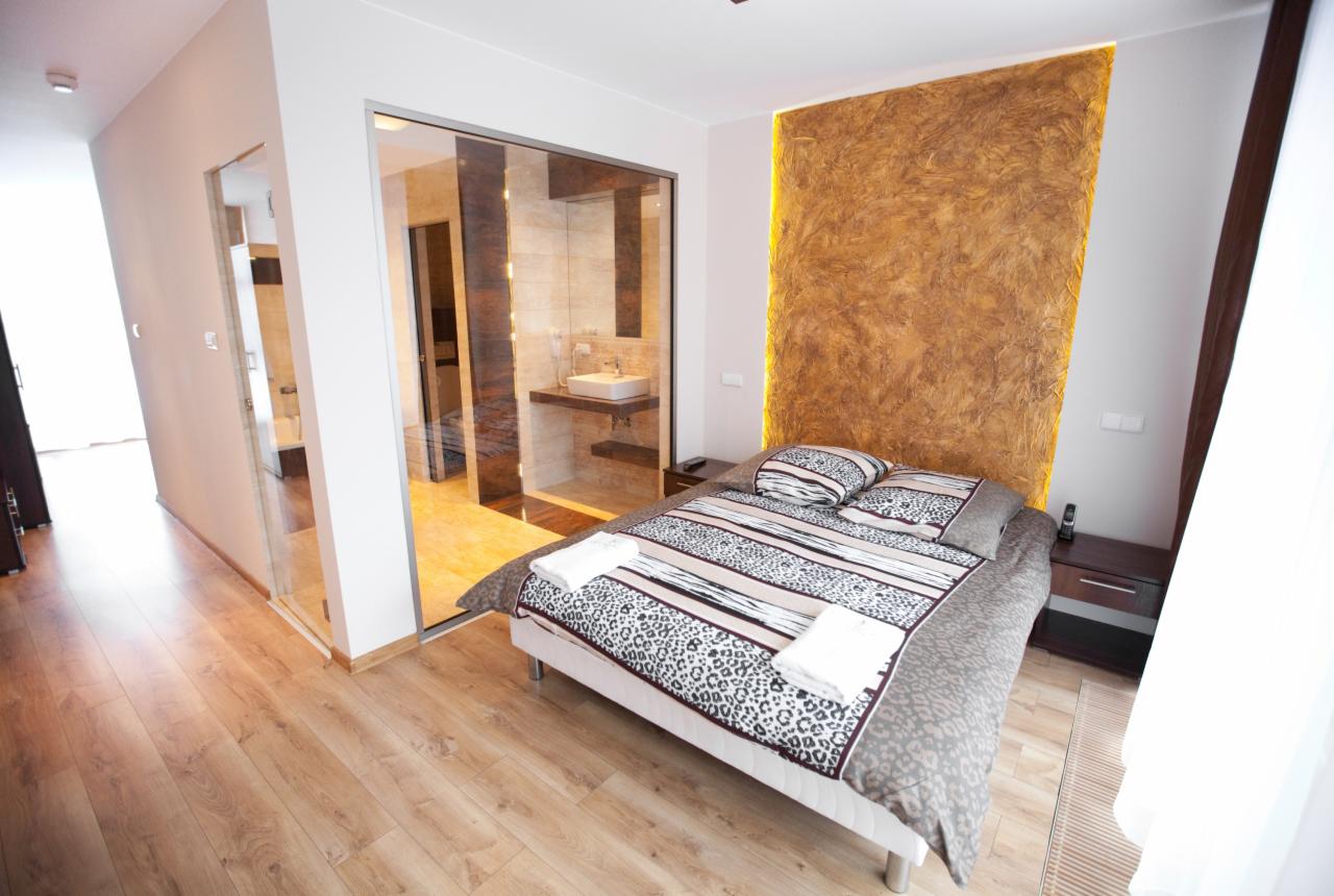 Pokój z łazienką oddzieloną szklaną ścianą, dekoracyjna ściana za łóżkiem z oświetleniem.