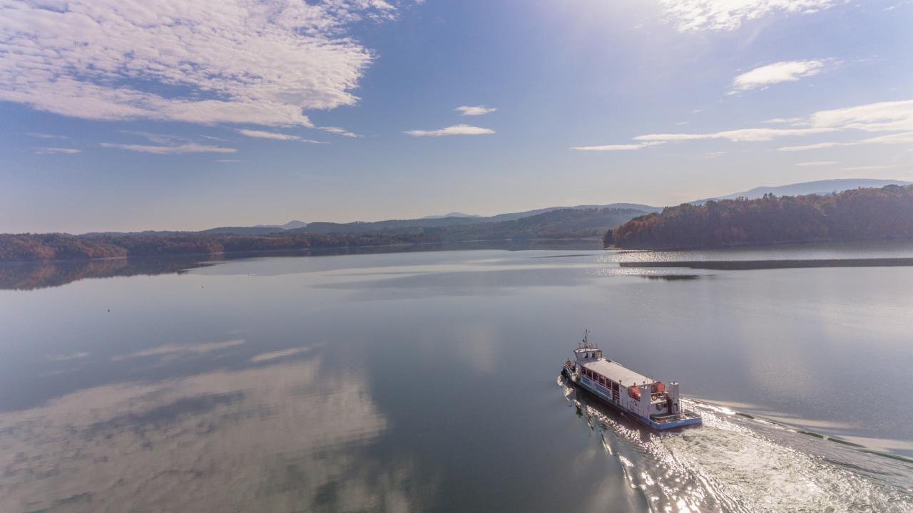 Prom pasażerski na spokojnym jeziorze z odbiciem chmur, w tle zalesione wzgórza w jesiennej kolorystyce, jasne słońce.
