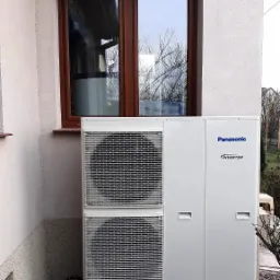Zewnętrzna jednostka grzewcza Panasonic Inverter zainstalowana przy ścianie domu z brązowym oknem, widoczna na betonowych podstawach i żwirowym podłożu.