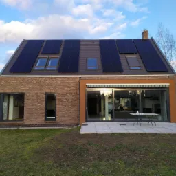 Nowoczesny dom jednorodzinny z brązowej cegły i pomarańczowego tynku z panelami fotowoltaicznymi na dachu i dużym tarasem z meblami ogrodowymi.