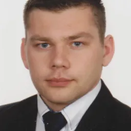 Portret młodego mężczyzny w ciemnym garniturze i niebieskim krawacie, na jasnym tle.