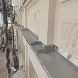 Zamontowana, błyszcząca, metalowa rynna na elewacji budynku z dekoracyjnymi elementami, widoczna z rusztowania.