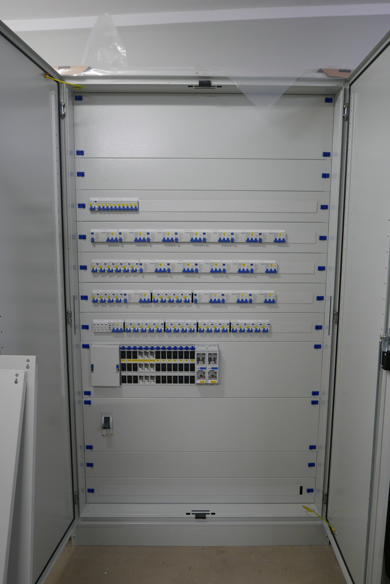 Otwarta rozdzielnia elektryczna w kolorze białym, z widocznymi bezpiecznikami, wyłącznikami różnicowoprądowymi i listwami zaciskowymi, zamontowana w nowym budynku, zabezpieczona folią przed...