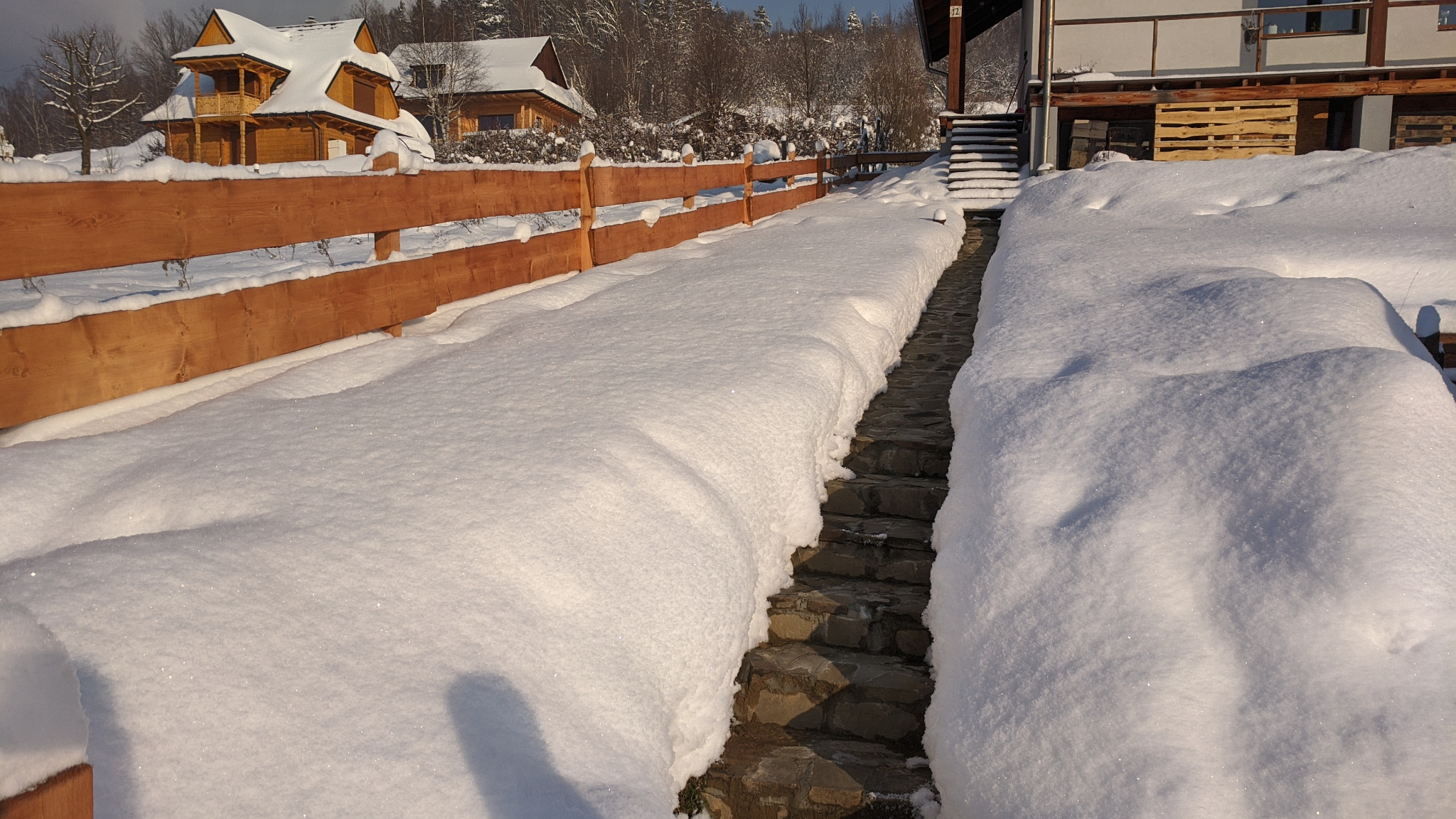 Zasypana śniegiem kamienna ścieżka prowadząca do budynku, widoczny drewniany płot i domy w tle, zimowy krajobraz.