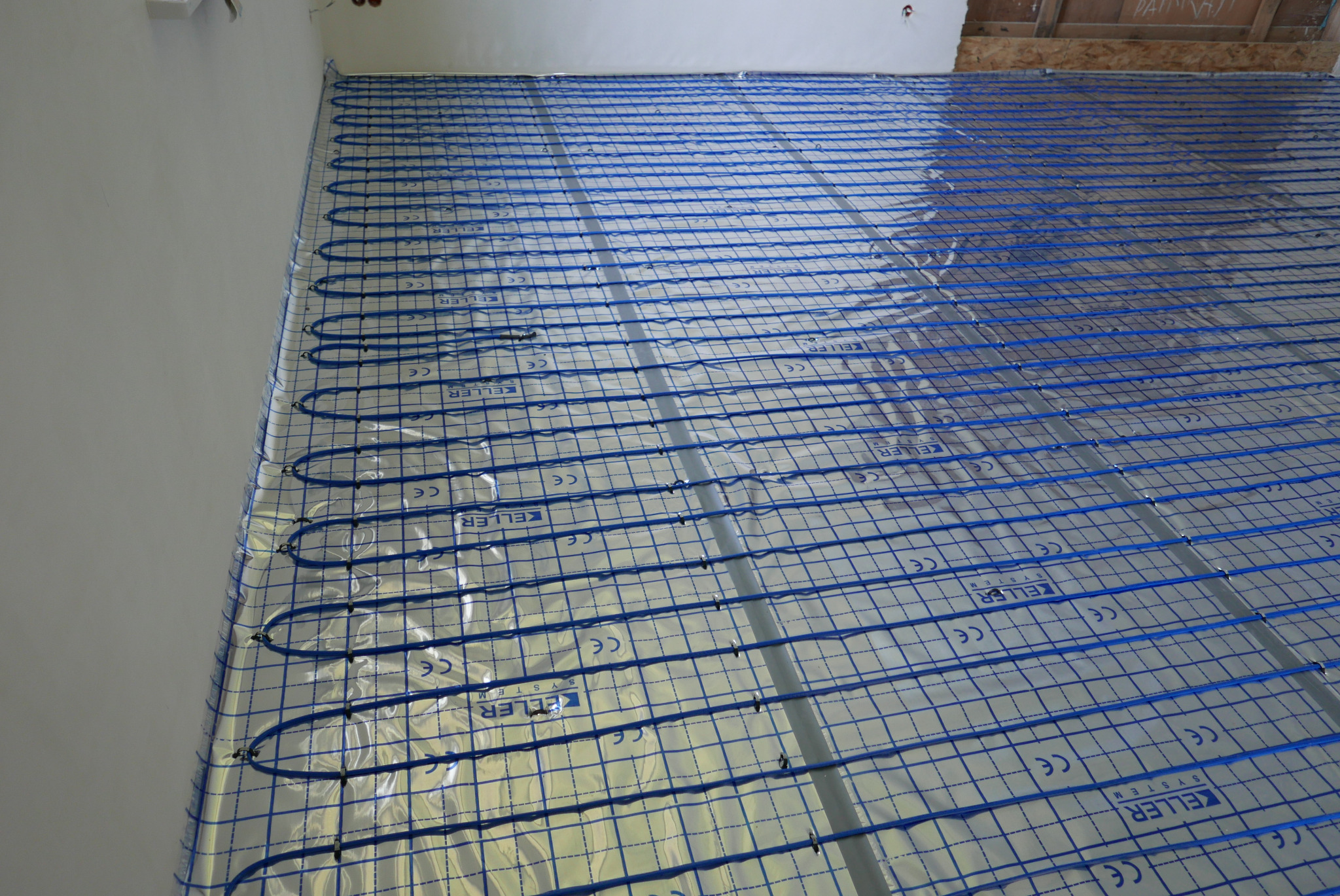 Niebieskie rurki elektrycznego systemu grzewczego ułożone na folii z nadrukowaną siatką i logo Keller System, przygotowane do montażu w nowym budynku.