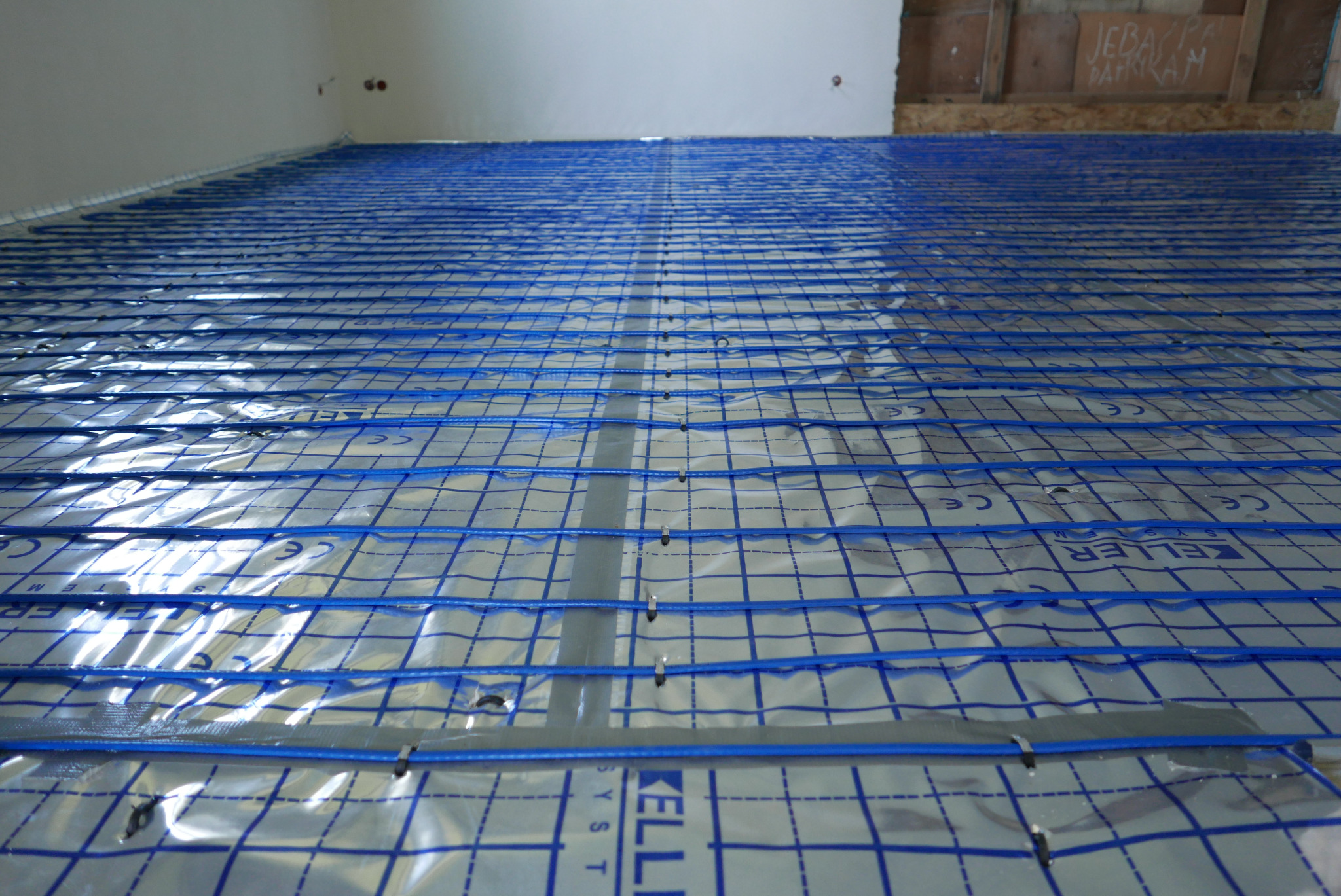Niebieskie przewody grzewcze ułożone równolegle na srebrnej folii z nadrukiem kratki, zamocowane taśmą i spinkami na podłodze w trakcie montażu systemu ogrzewania podłogowego.