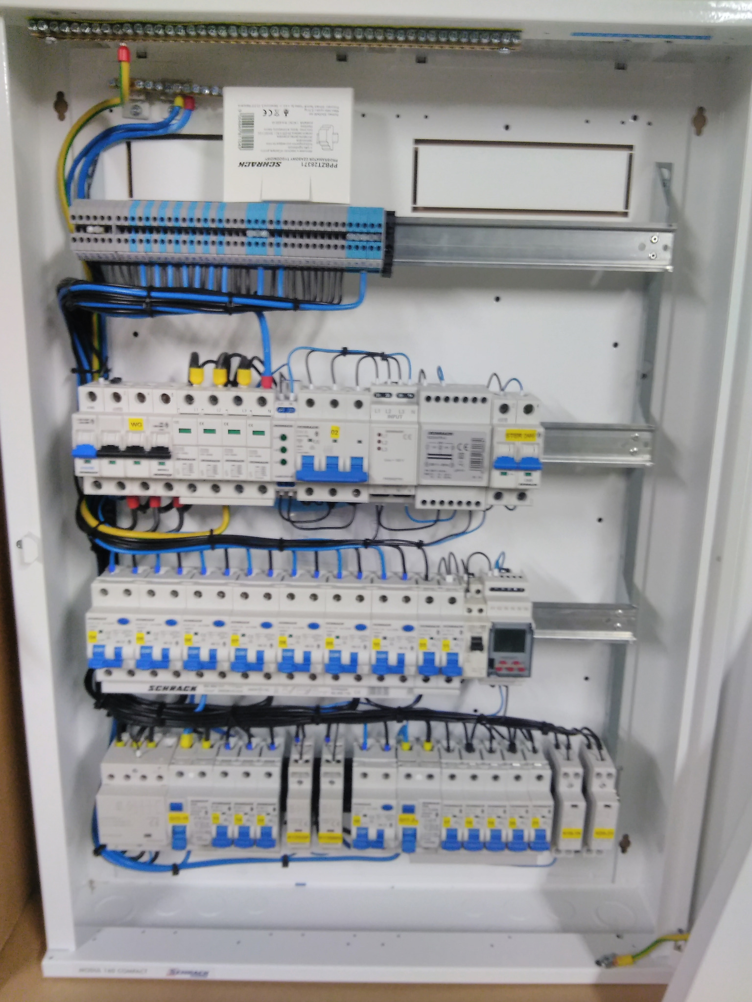 Wnętrze białej rozdzielnicy elektrycznej marki Schrack, z widocznymi bezpiecznikami, przewodami w kolorach niebieskim, żółtym i czarnym oraz listwami zaciskowymi.