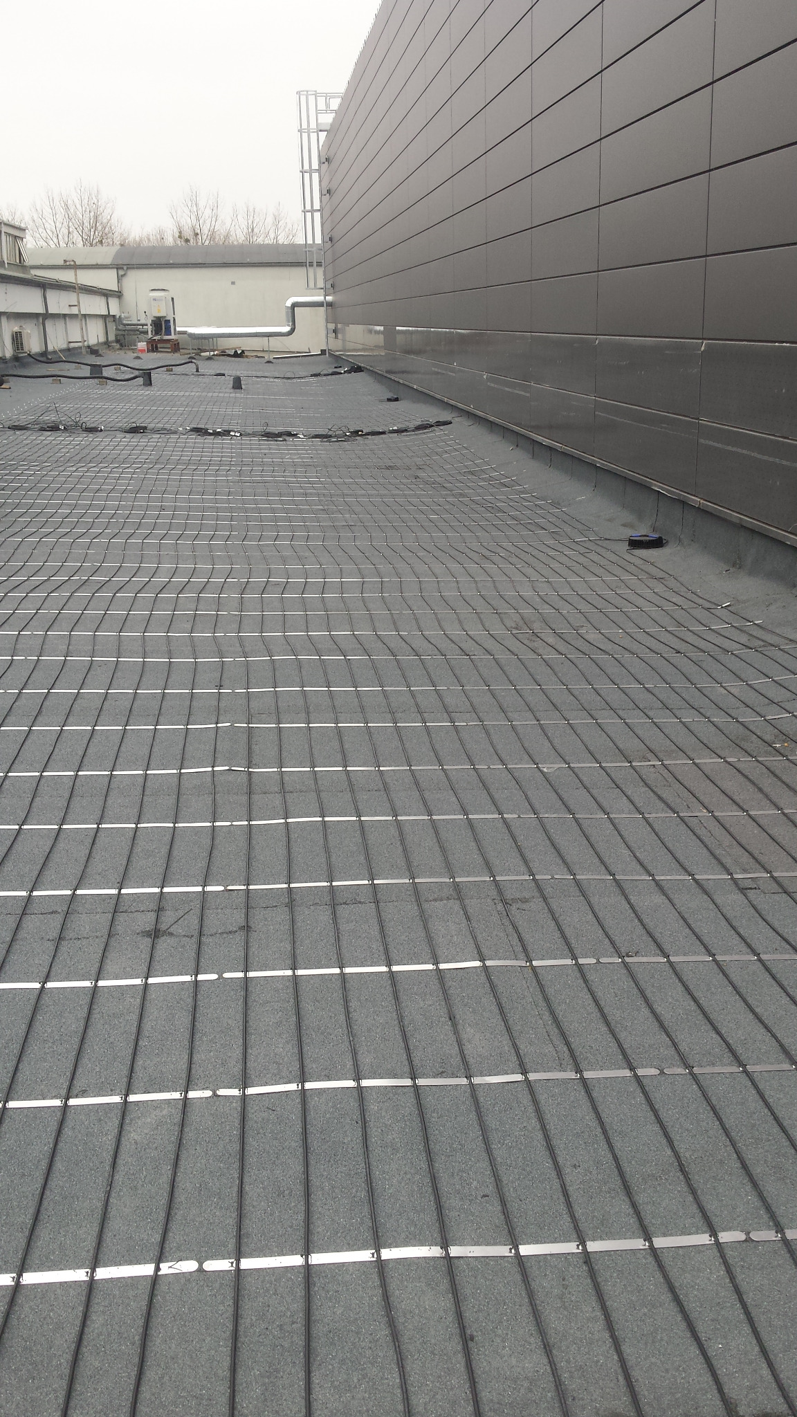Montaż mat grzewczych na dachu budynku przemysłowego, widoczne równoległe kable grzewcze z metalowymi mocowaniami, szara membrana dachowa, budynek w tle.