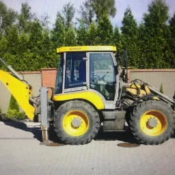 Żółto-szara koparko-ładowarka zaparkowana na kostce brukowej, z widocznym przednim i tylnym ramieniem, tło stanowi mur z cegły i wysokie tuje.