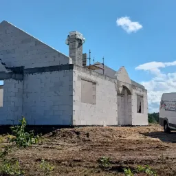 Wznoszenie ścian domu z betonu komórkowego, widoczny komin w trakcie budowy oraz firmowy bus z reklamą na tle błękitnego nieba z chmurami.