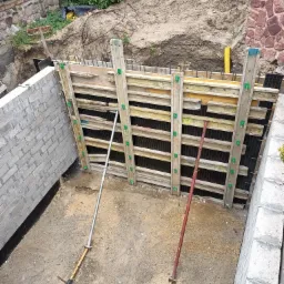 Szczegółowe ujęcie szalunku fundamentowego z deskami i metalowymi rozpórkami, widoczny fragment ściany z bloczków betonowych oraz wykop ziemny w tle.
