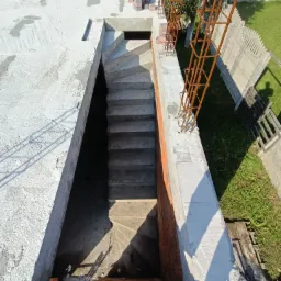 Wykop schodowy w surowym stanie, z widocznymi stopniami i zbrojeniami, betonowe ściany szalunkowe, widok z góry na buty robotnika.