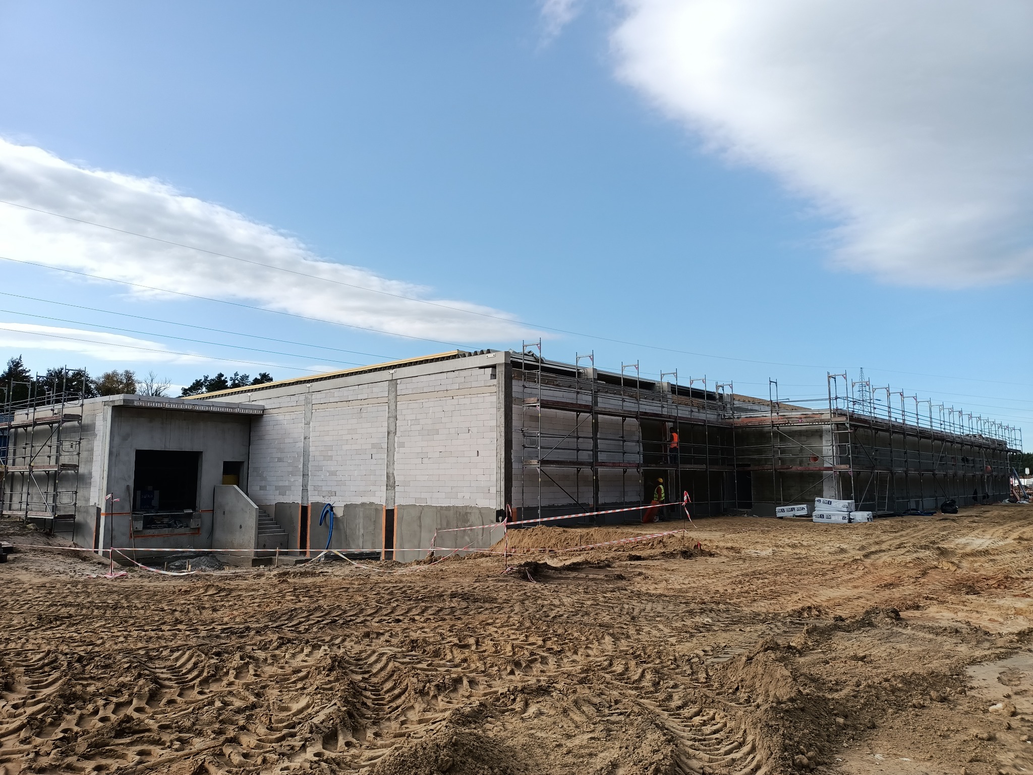 Budynek w stanie surowym otwartym z białego bloczka, otoczony rusztowaniami, widoczny pracownik w pomarańczowej kamizelce, teren wokół budynku z wykopaną ziemią i śladami opon.