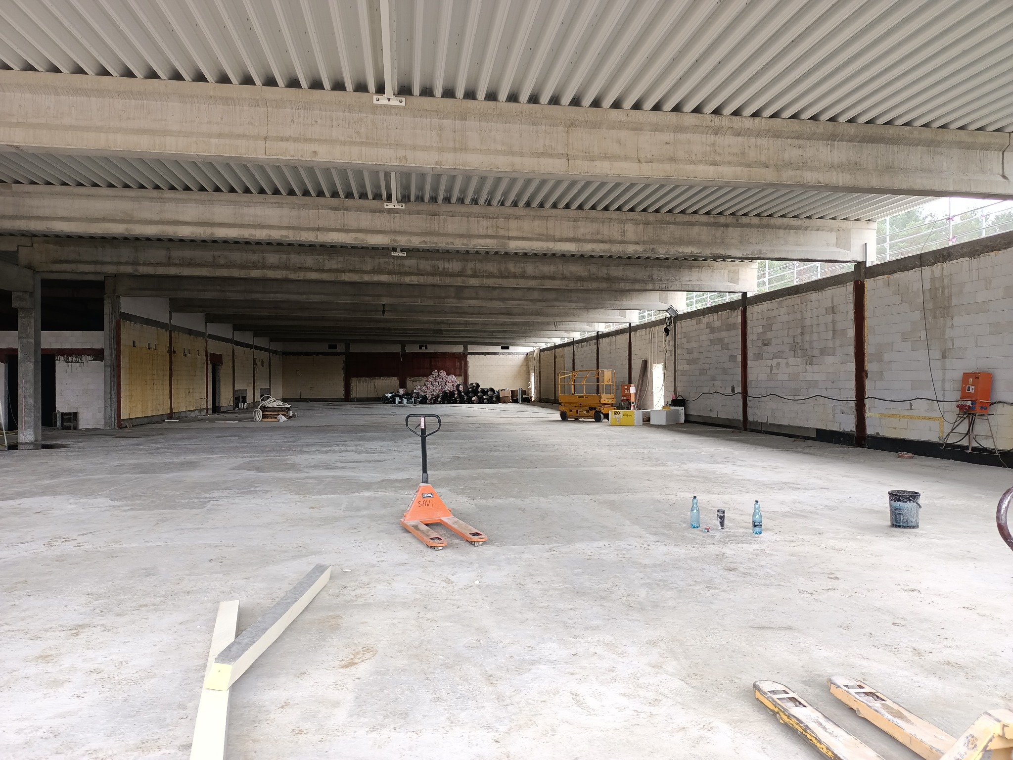 Wnętrze hali w trakcie budowy, widoczne ściany z bloczków, betonowy strop, wózek paletowy na posadzce, butelki z wodą i narzędzia budowlane.