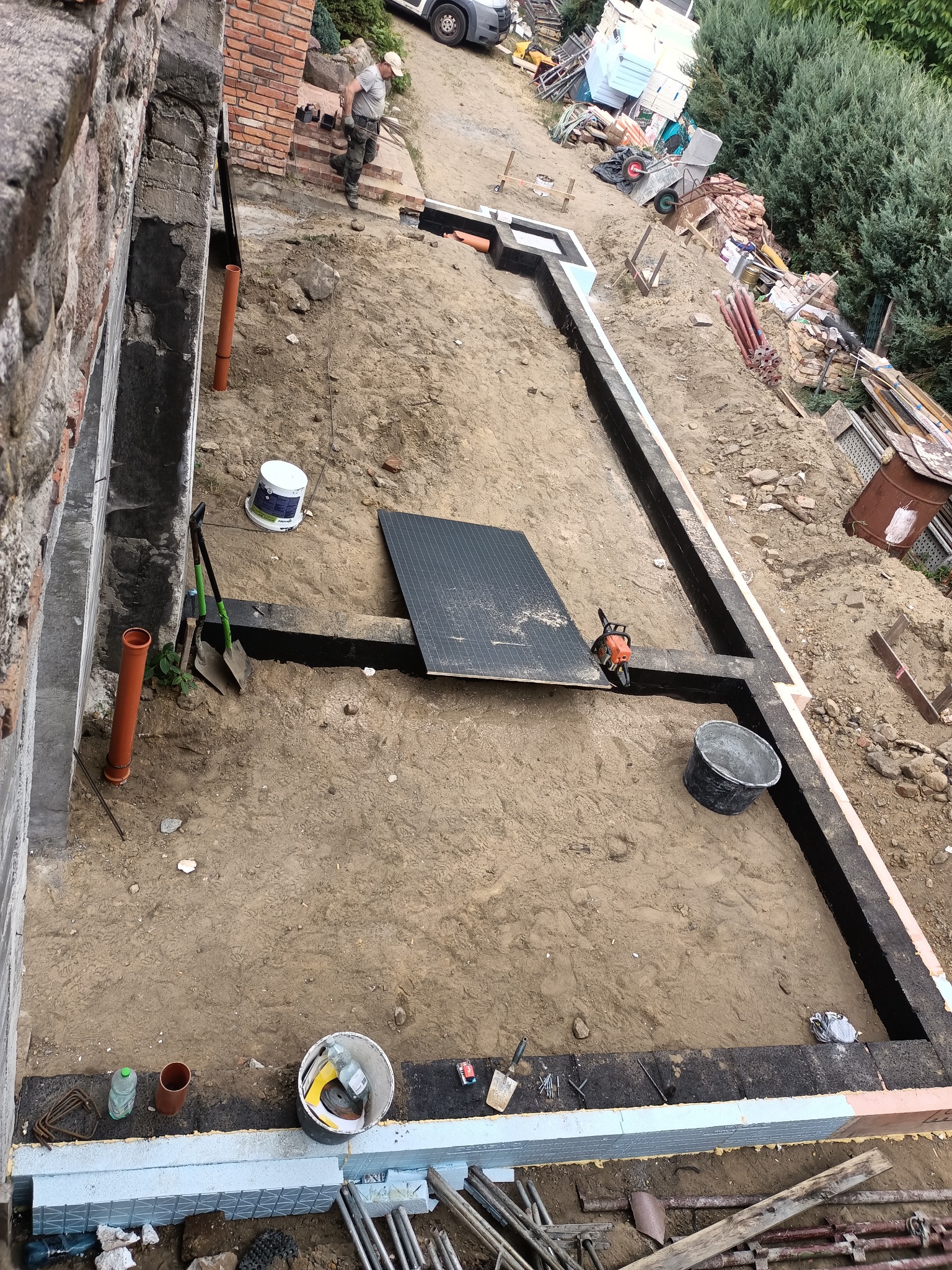 Budowa fundamentów: widok z góry na wykop z izolacją termiczną, narzędzia budowlane i pracujący robotnik w tle.