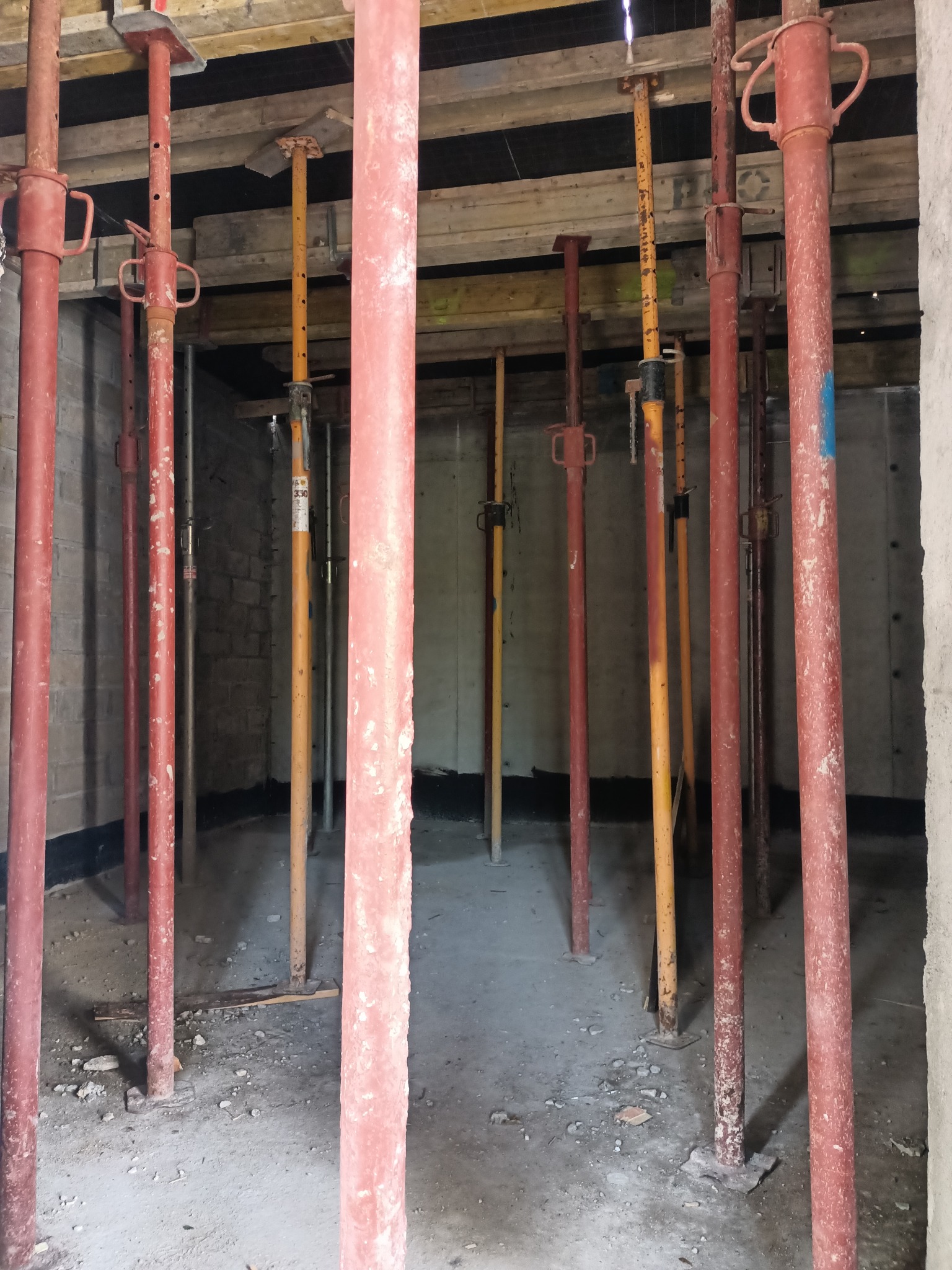 Wnętrze surowego budynku z ceglanymi ścianami i betonową podłogą, podparte licznymi, czerwonymi i żółtymi, stalowymi stemplami budowlanymi, podtrzymującymi drewniany szalunek stropu.