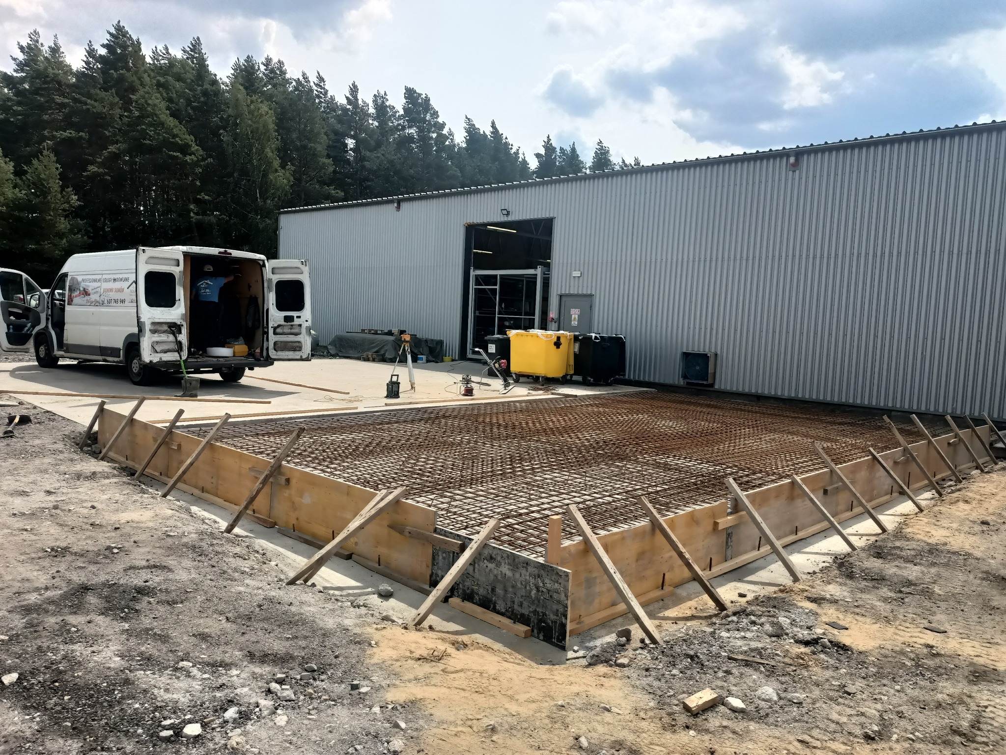 Przygotowanie zbrojenia i szalunku pod płytę fundamentową na gruncie, widoczny samochód dostawczy i budynek magazynowy w tle.