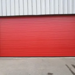 Czerwona, pozioma brama garażowa segmentowa z brązową ramą, zamontowana w szarym budynku z blachy falistej.