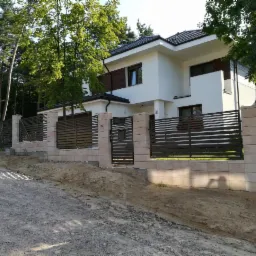 Nowoczesna posesja z ogrodzeniem panelowym z poziomymi przęsłami w kolorze antracytowym i słupkami z jasnego kamienia, widoczny dwukondygnacyjny dom z białymi ścianami i ciemną dachówką, teren...