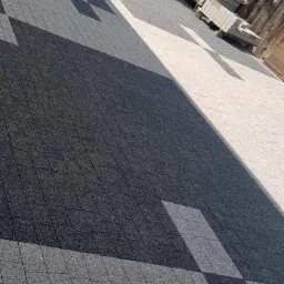 Ułożona nawierzchnia z kostki granitowej w odcieniach szarości i bieli, tworząca geometryczny wzór. W tle widoczne palety z kostką i drewniane ogrodzenie.