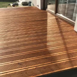 Nowoczesny taras z desek o ciepłym, brązowym odcieniu, z widocznymi drobnymi śladami po montażu i odbiciem światła w szklanych drzwiach przesuwnych domu.