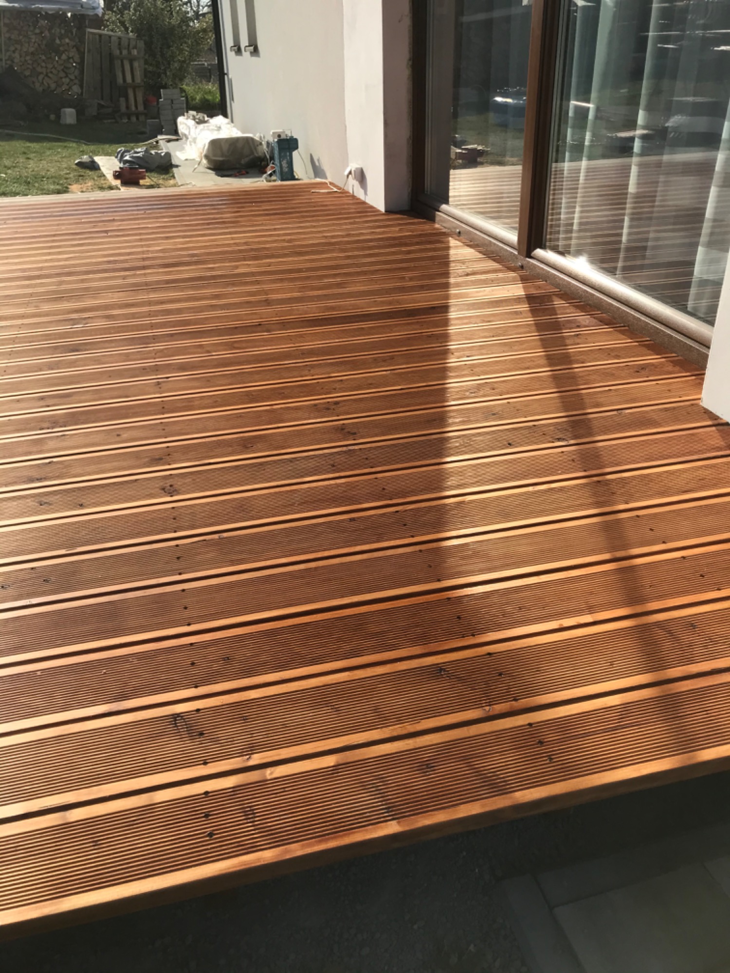 Nowoczesny taras z desek o ciepłym, brązowym odcieniu, z widocznymi drobnymi śladami po montażu i odbiciem światła w szklanych drzwiach przesuwnych domu.