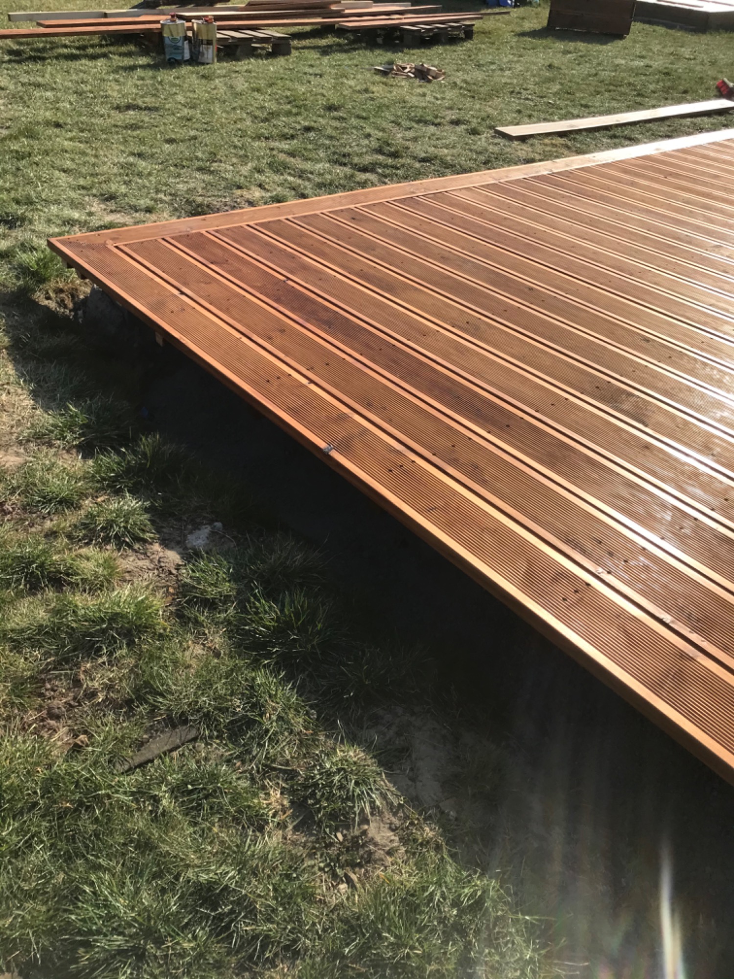 Fragment nowo wybudowanego tarasu z desek kompozytowych, widoczne deski ułożone równolegle, trawa w tle i materiały budowlane.