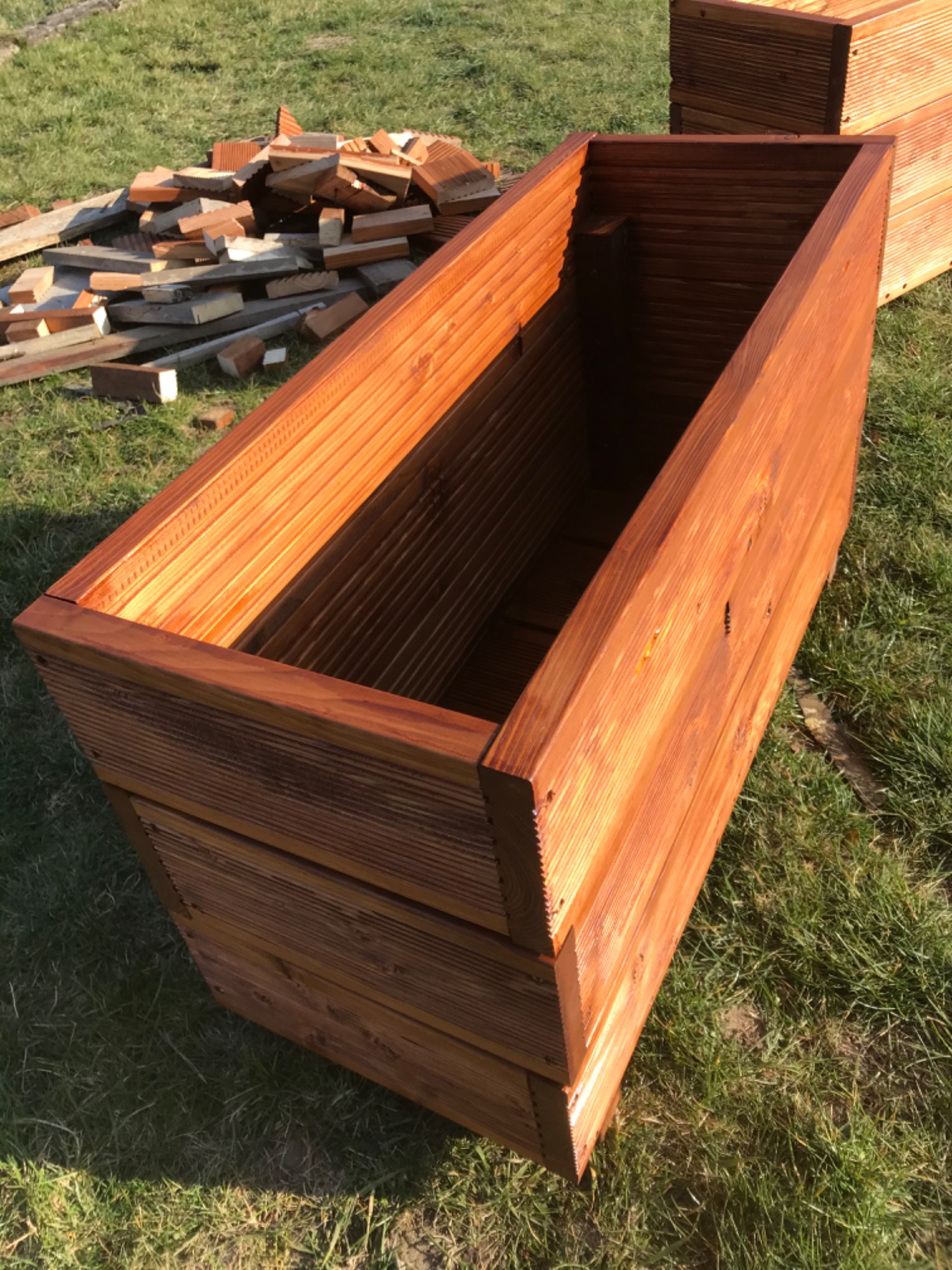 Dwie drewniane skrzynie na rośliny, wykonane z desek, pomalowane na brązowo, stoją na trawie obok sterty cegieł i desek.