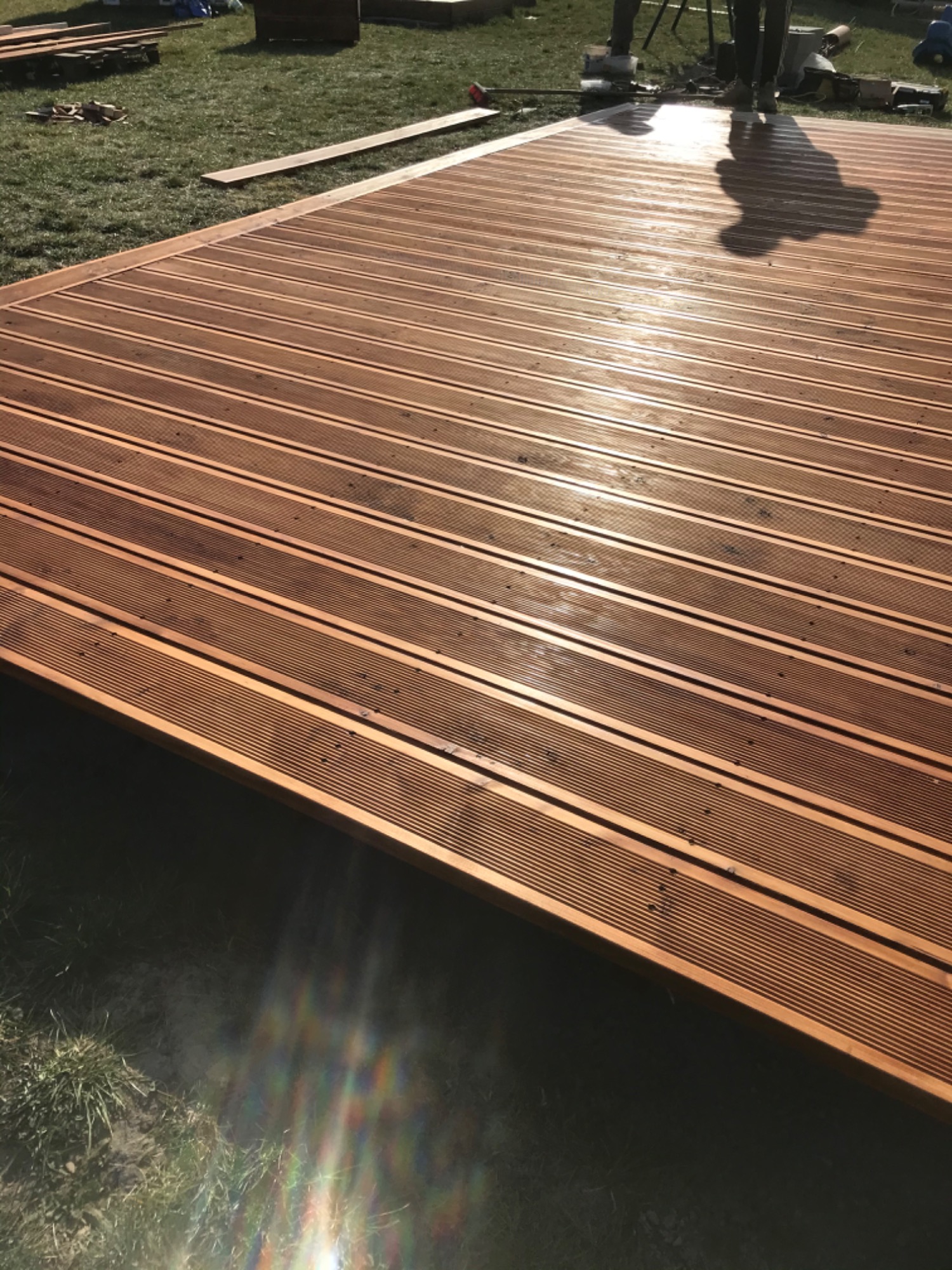 Nowo wybudowany taras z desek ryflowanych, częściowo ukończony, z widocznym cieniem osoby stojącej na nim i deskami przygotowanymi do dalszego montażu na trawniku.