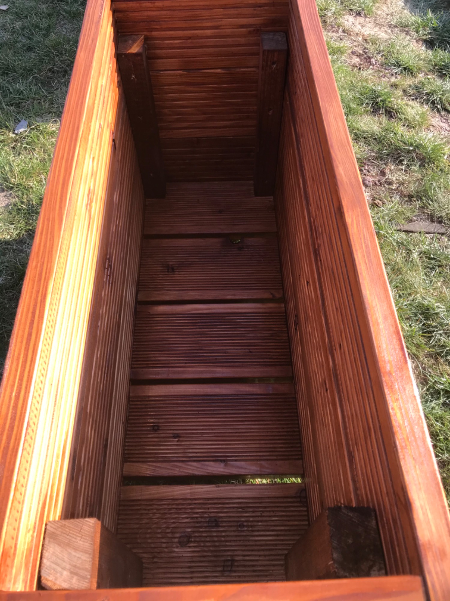 Wnętrze pustej, brązowej, drewnianej skrzyni na rośliny, wykonanej z desek, widoczne z góry na tle trawy.