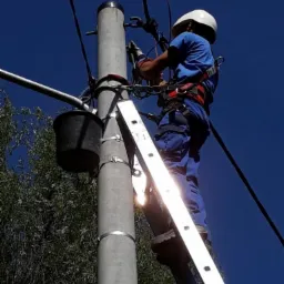 Elektryk w kasku i uprzęży na drabinie, pracuje przy słupie energetycznym z kablem, na tle błękitnego nieba i zieleni drzew.