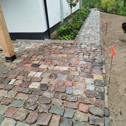 Hydro-Tech Katarzyna Rudź - Ścieżka z kostki granitowej w odcieniach szarości, brązu i czerwieni, prowadząca wzdłuż budynku z białymi ścianami i rabatami kwiatowymi, z widocznymi pomarańczowymi znacznikami wzdłuż krawędzi...