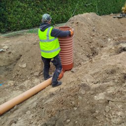 Hydro-Tech Katarzyna Rudź - Pracownik w kamizelce odblaskowej montuje brązowy zbiornik z tworzywa sztucznego w wykopie ziemnym, obok leży rura, w tle żywopłot i koparka.