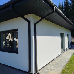 Hydro-Tech Katarzyna Rudź - Nowoczesny dom jednorodzinny z białą elewacją, czarnym dachem i rynnami. Kostka brukowa przed budynkiem i trawnik. Widok z perspektywy.