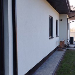 Hydro-Tech Katarzyna Rudź - Elewacja o jasnej strukturze z ciemnymi oknami i rynnami. Widoczny fragment chodnika z kostki brukowej i trawnik. Dom z zewnątrz po remoncie.