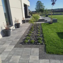 Nowoczesny ogród z szarą kostką brukową, rabatą z kamieni i zielenią, donice przy elewacji budynku. Starannie zaprojektowana przestrzeń.