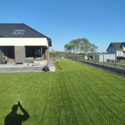 Hydro-Tech Katarzyna Rudź - Nowoczesny ogród z idealnie przystrzyżonym trawnikiem, niskimi krzewami i ogrodzeniem panelowym. W tle dom z tarasem i meblami ogrodowymi.