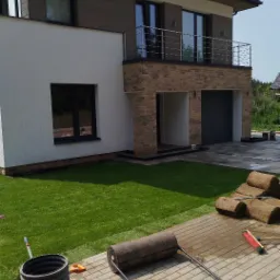 Nowoczesny dom z równo ułożoną trawą z rolki, wałem do trawy i paletą kostki brukowej. Widoczne detale jak ceglana elewacja i balkon.