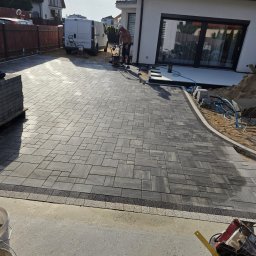 Hydro-Tech Katarzyna Rudź - Ułożona kostka brukowa w szarych odcieniach, z obrzeżem z drobnej kostki. Pracownik zagęszcza teren. W tle biały budynek i zaparkowany bus.