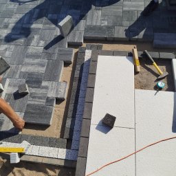 Hydro-Tech Katarzyna Rudź - Fragment układania kostki brukowej z różnych materiałów i kolorów. Częściowo ułożona nawierzchnia z narzędziami pracy. Widoczny etap prac.