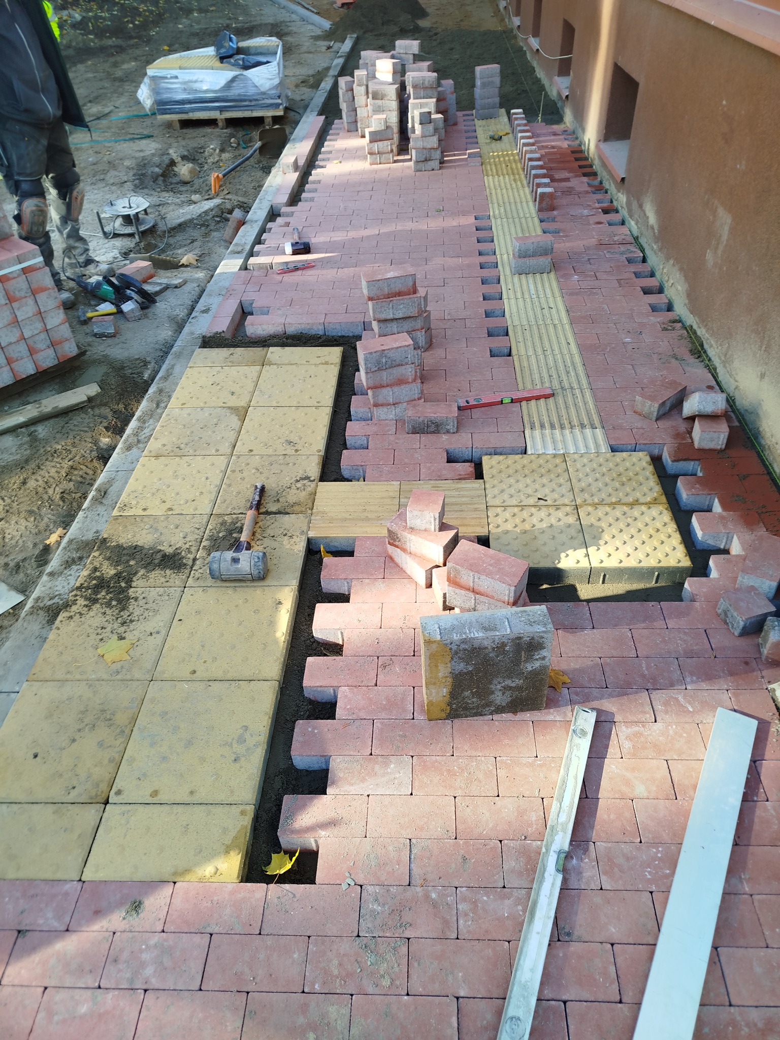 Fragment chodnika w trakcie budowy z czerwonej kostki brukowej i żółtych płyt z wypustkami, widoczne narzędzia budowlane, poziomica i gumowy młotek, paleta kostki w tle.