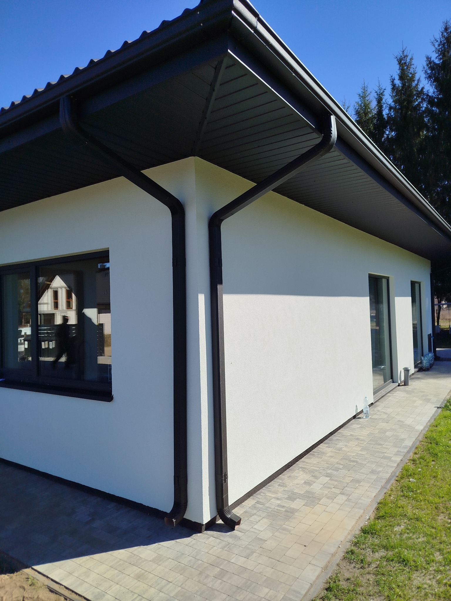 Nowoczesny dom jednorodzinny z białą elewacją, czarnym dachem i rynnami. Kostka brukowa przed budynkiem i trawnik. Widok z perspektywy.