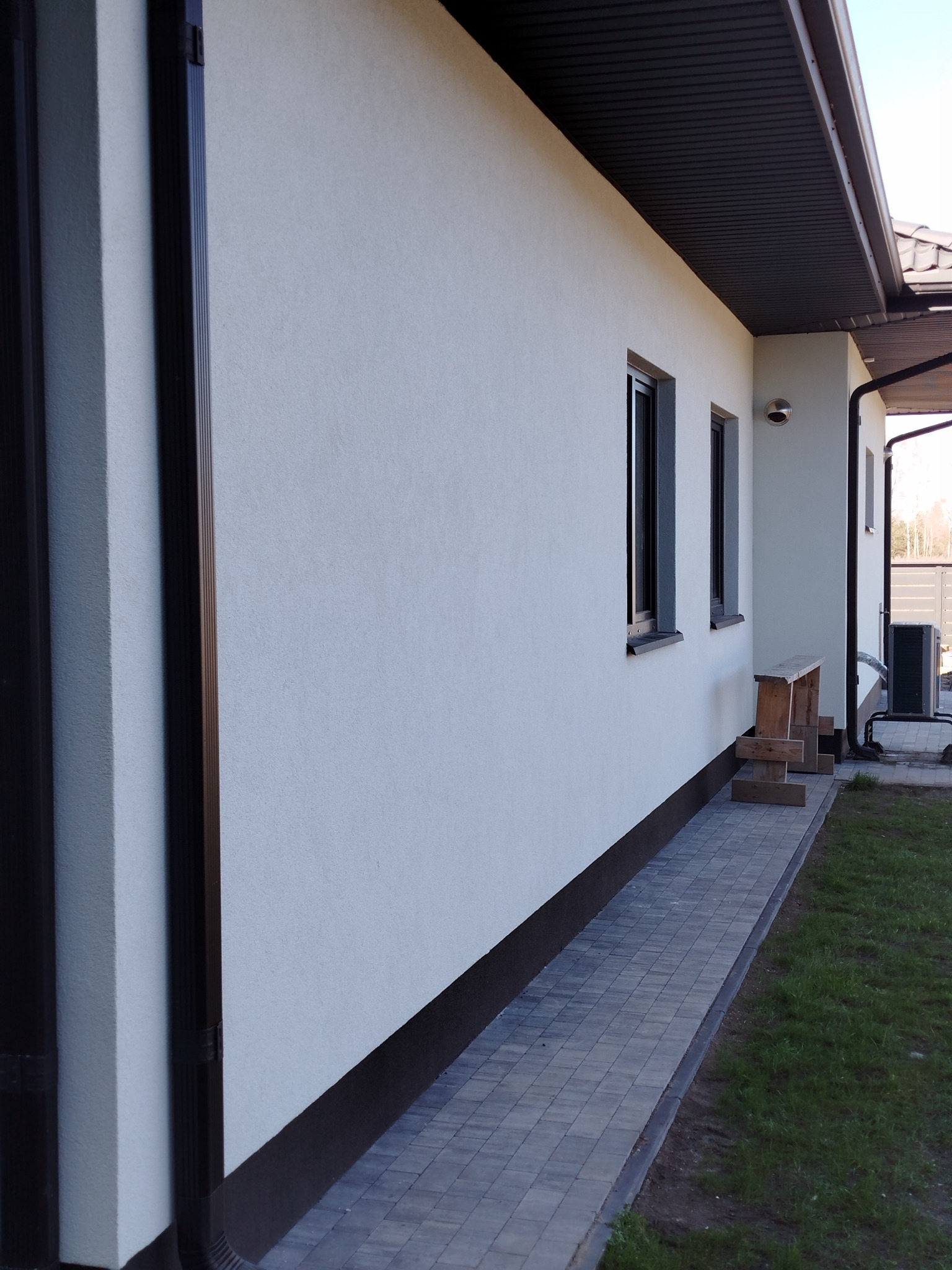 Elewacja o jasnej strukturze z ciemnymi oknami i rynnami. Widoczny fragment chodnika z kostki brukowej i trawnik. Dom z zewnątrz po remoncie.