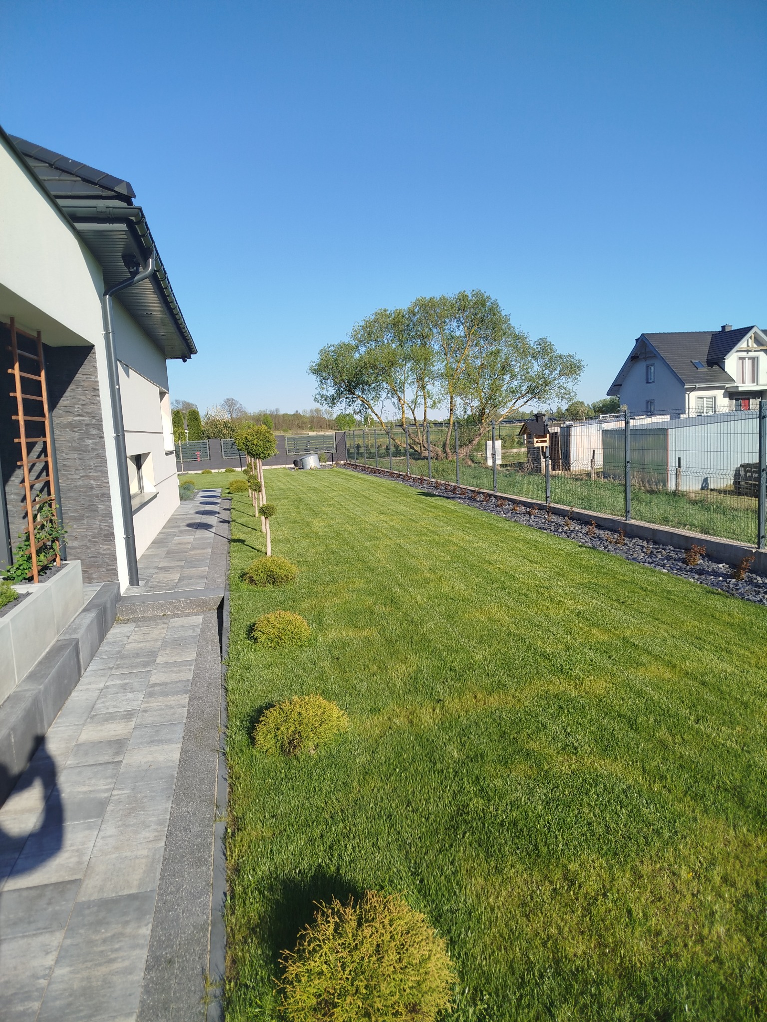 Zadbany trawnik z kostką brukową i nasadzeniami wzdłuż ogrodzenia panelowego. Widok na dom z szarą elewacją i elementami drewnianymi.