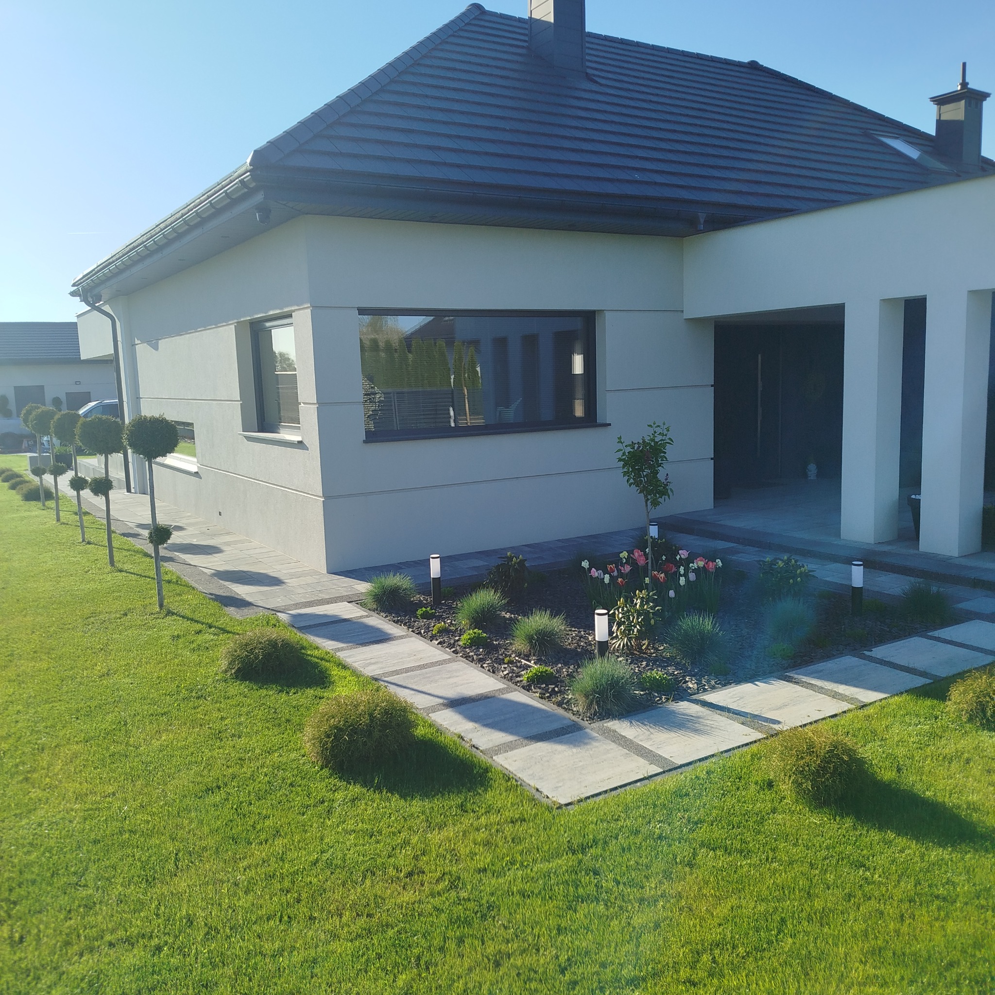 Nowoczesny dom z zadbanym ogrodem: trawnik, rabaty kwiatowe, ścieżka z kostki i formowane drzewka. Oświetlenie rabat i elewacja w jasnych barwach.