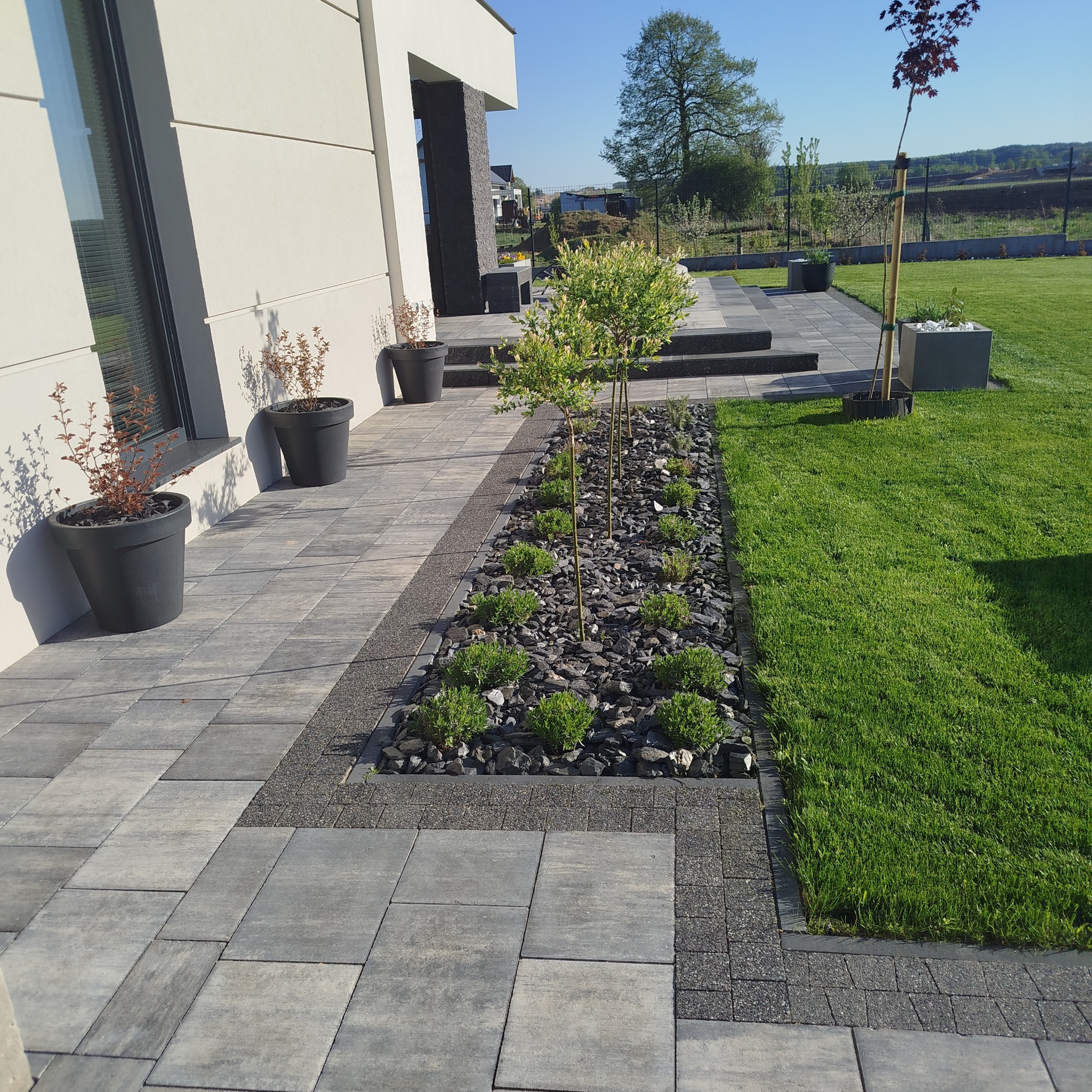 Nowoczesny ogród z szarą kostką brukową, rabatą z kamieni i zielenią, donice przy elewacji budynku. Starannie zaprojektowana przestrzeń.